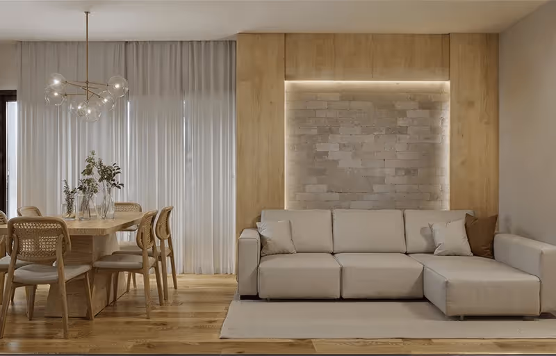 Sala de estar moderna com sofá bege em forma de L e área de jantar com mesa e cadeiras de madeira ao lado de uma janela com cortinas brancas.