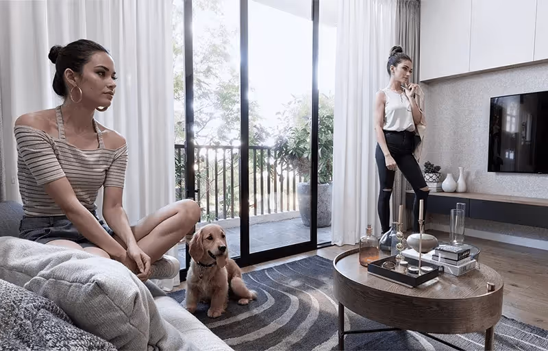 Duas mulheres em uma sala moderna, uma sentada no sofá e outra em pé perto da mesa de centro, com um cachorro ao lado da mulher sentada.