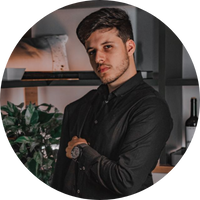 Homem jovem com camisa preta e relógio, posando com os braços cruzados em frente a uma prateleira com plantas e garrafas.