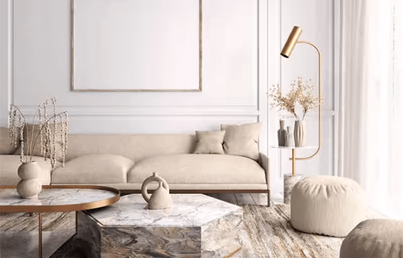 Sala de estar minimalista com sofá bege, almofadas, mesa de centro com tampo de mármore, puff bege, vaso com flores secas e luminária dourada.