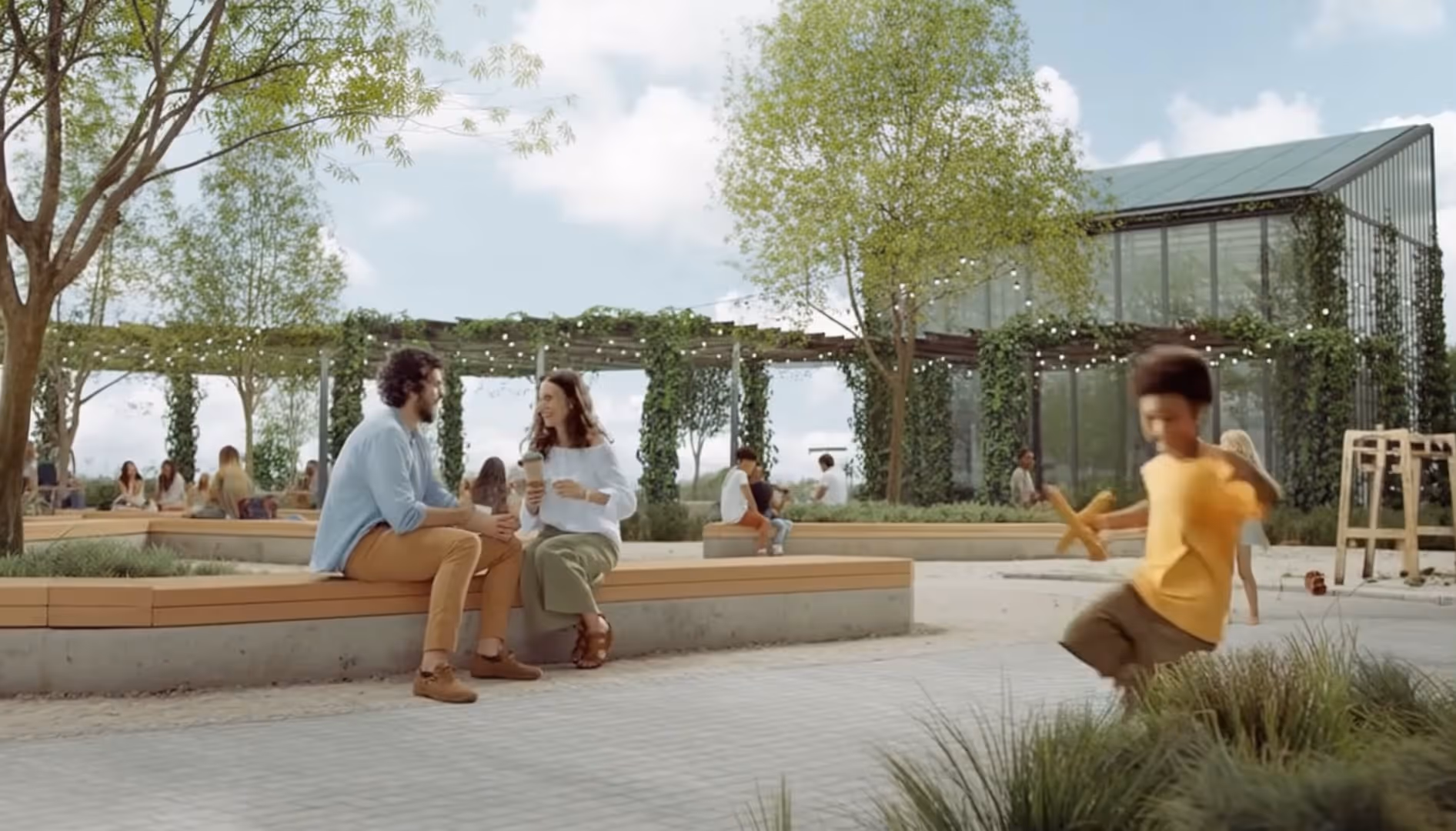 Casal conversando e tomando café em banco ao ar livre em um parque com crianças brincando e estrutura de vidro coberta por plantas ao fundo.