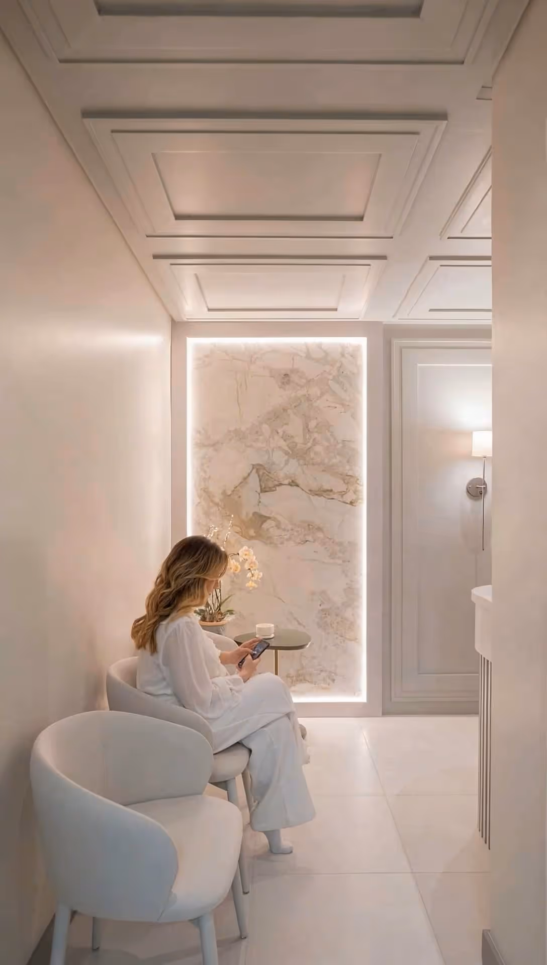 Mulher sentada em cadeira branca em sala minimalista com parede decorada de mármore e mesa lateral.