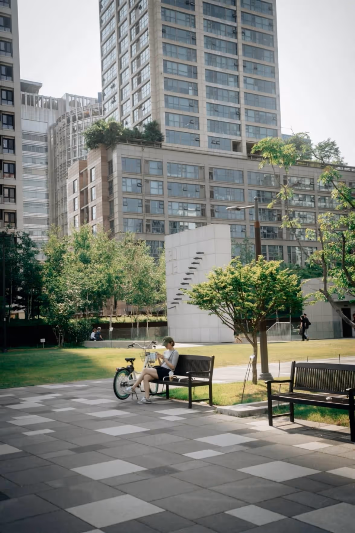 Pessoa sentada em um banco de parque ao lado de uma bicicleta em área urbana com árvores e prédios ao redor.